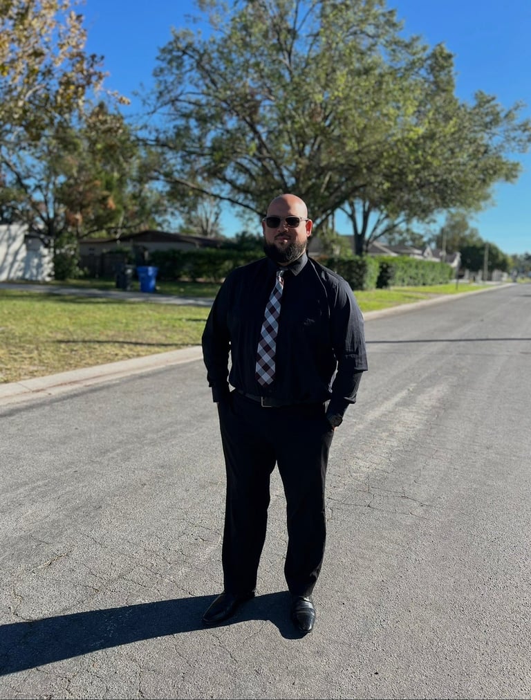 Man in black business suit with checkered tie and sunglasses standing on a residential street under trees on a sunny day