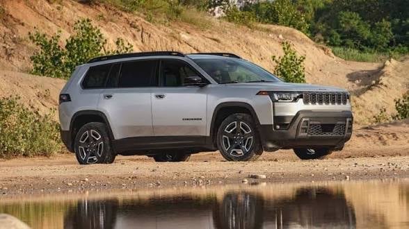 Silver Jeep Wagoneer parked on gravel beside water with rocky hillside in background