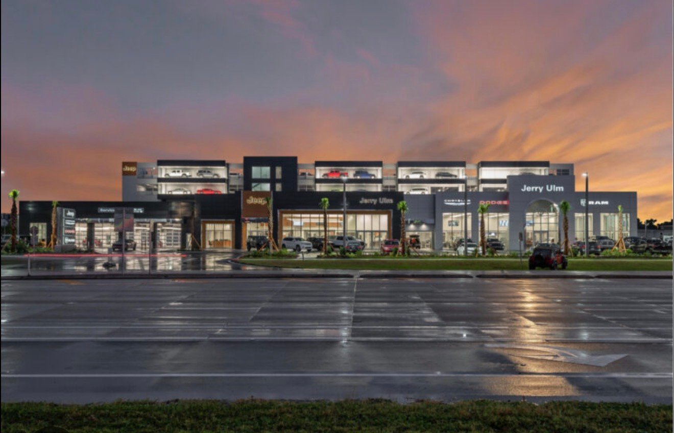 Modern retail building with illuminated storefront at dusk, reflecting on wet parking lot under colorful sunset sky