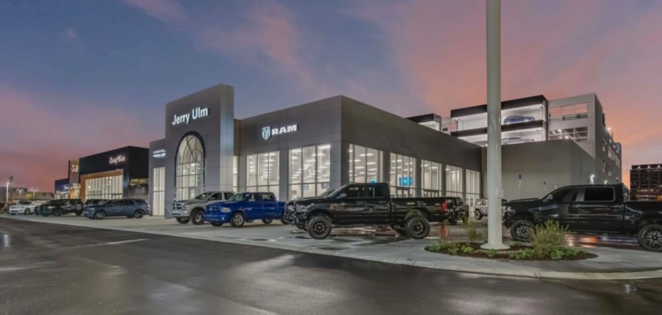 Modern automotive dealership showroom with glass facade and blue vehicles displayed outside during dusk with dramatic sky